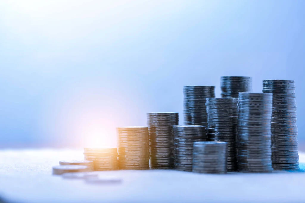 Piles of coins of different heights on a blue background with light shining on the two of the piles 5 inventory reduction methods to deliver cost savings