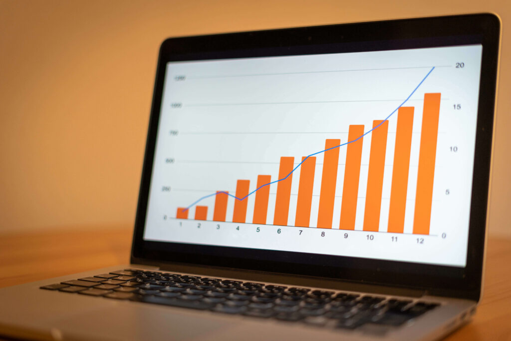 A close-up of a laptop with the screen showing a bar chart with orange bars, with a blue line chart over the top. The orange block shows an increasing trend, and the blue line is around the middle of the orange block but ends above it. The background is two shades of orange. How to calculate forecast accuracy and forecast error.
