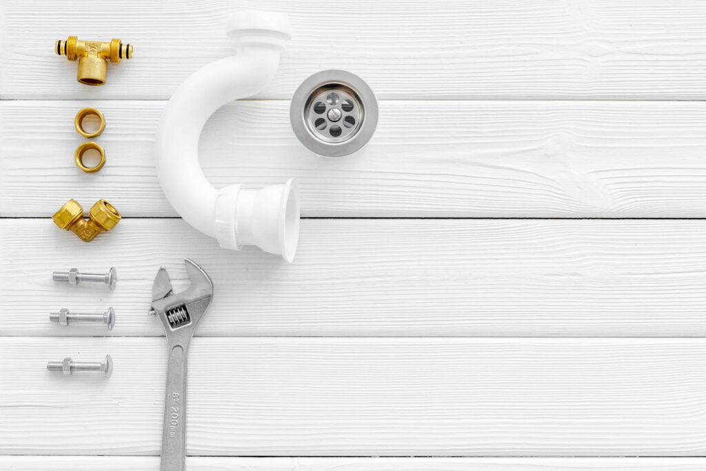 A white wooden background with a white u-bend and some gold and silver plumbing fixtures, a silver plug drain and a silver spanner to show bill of materials