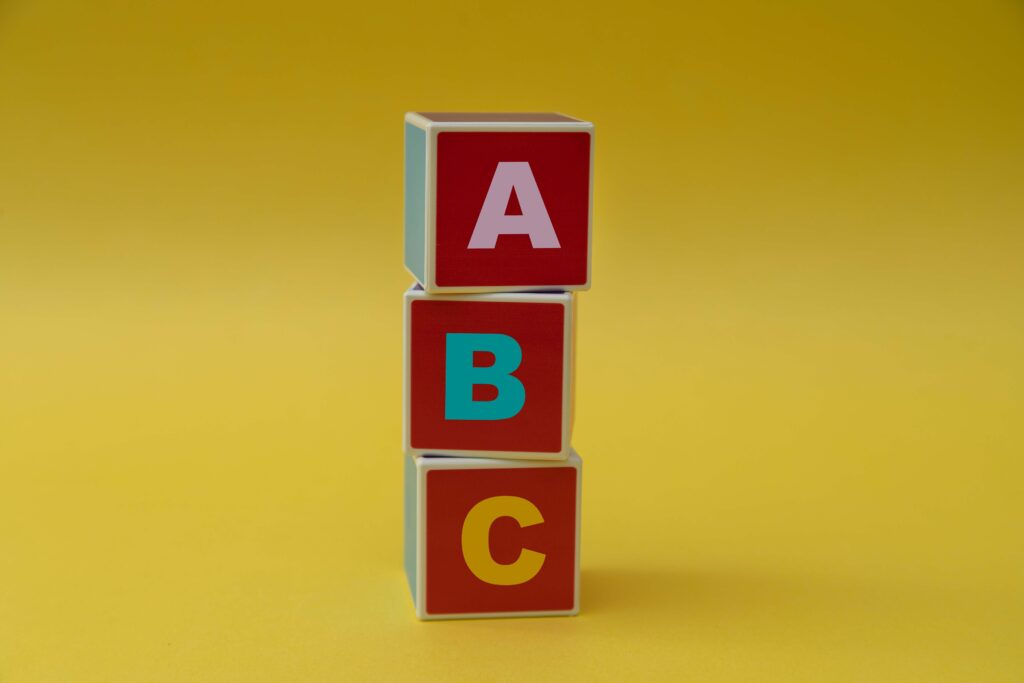 A yellow background with three red, wooden blocks stacked on top of each other haphazardly the top one has a pink A, the middle one has a pale blue B and the bottom one has a yellow c ABC analysis in stock management