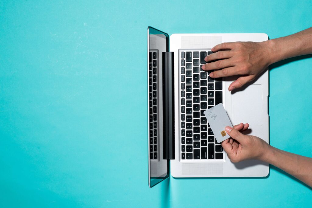 A person using a laptop to buy something online holding a credit card in their left hand, typing with their right hand, on a light blue background. The laptop is at a right angle to the bottom on the image. Supply chain management for ecommerce success