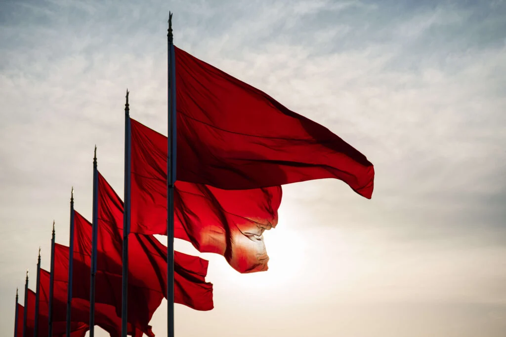A row of red flags blowing in the wind with a sun set in the background with white clouds and some blue patches to show warnings