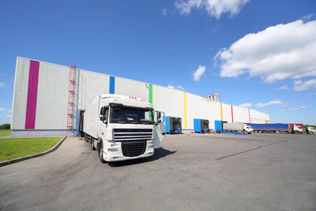 A large white truck reversed up to a row of white warehouses that have a coloured vertical stripe down the front of pink, blue, green, yellow, orange and purple. There are more lorries along the line and people at the warehouse doors. Supply chain disruption.
