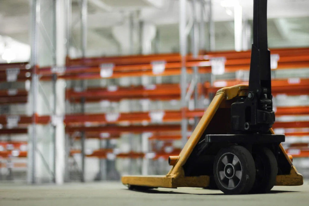Manual hydraulic lift in the foreground of an image of warehouse with shelving units behind slightly blurry
