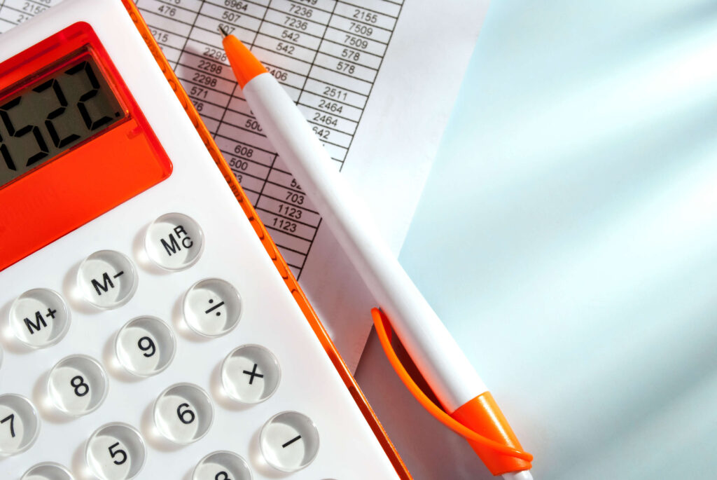 A white calculator with an orange frame around the screen next to a white pen with an orange surround around the tip and the top on a spreadsheet