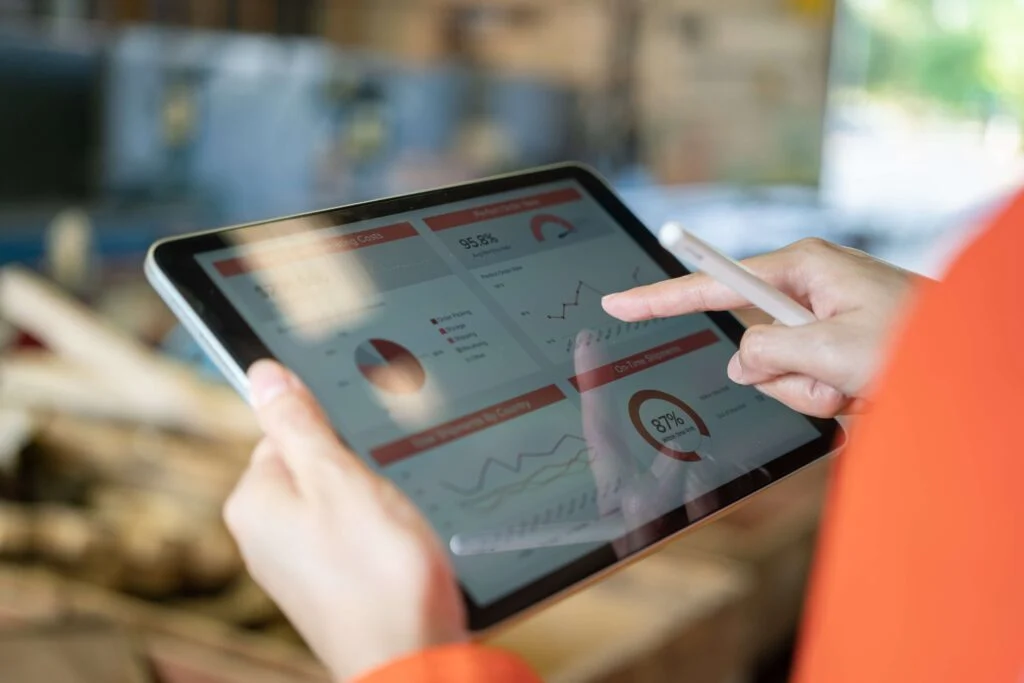 A person wearing an orange jumper, holding an iPad horizontally, you can only see the left shoulder and hands. The iPad has charts and data on and in the background is a blurry warehouse