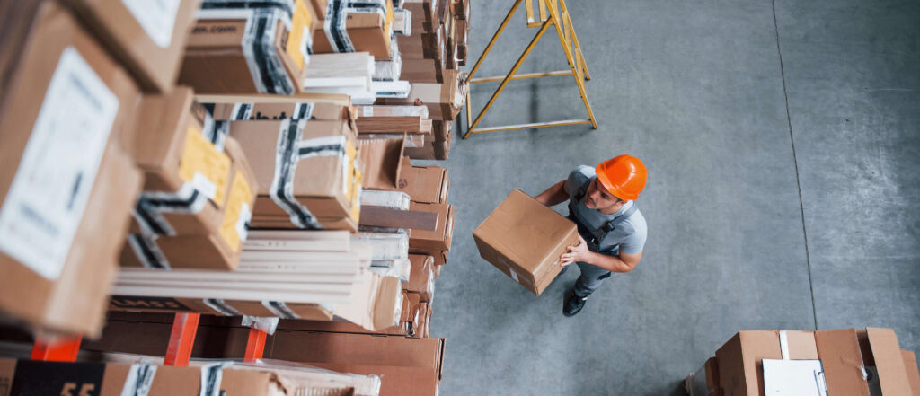Man in an orange hard hat wearing a grey t-short and blue dungarees holding a box in a warehouse, looking up at a more boxes stacked on shelves.