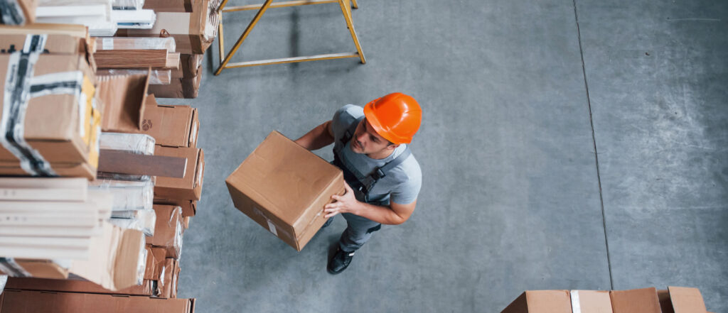 Man in an orange hard hat holding a box in a warehouse next to a shelving unit full of boxes